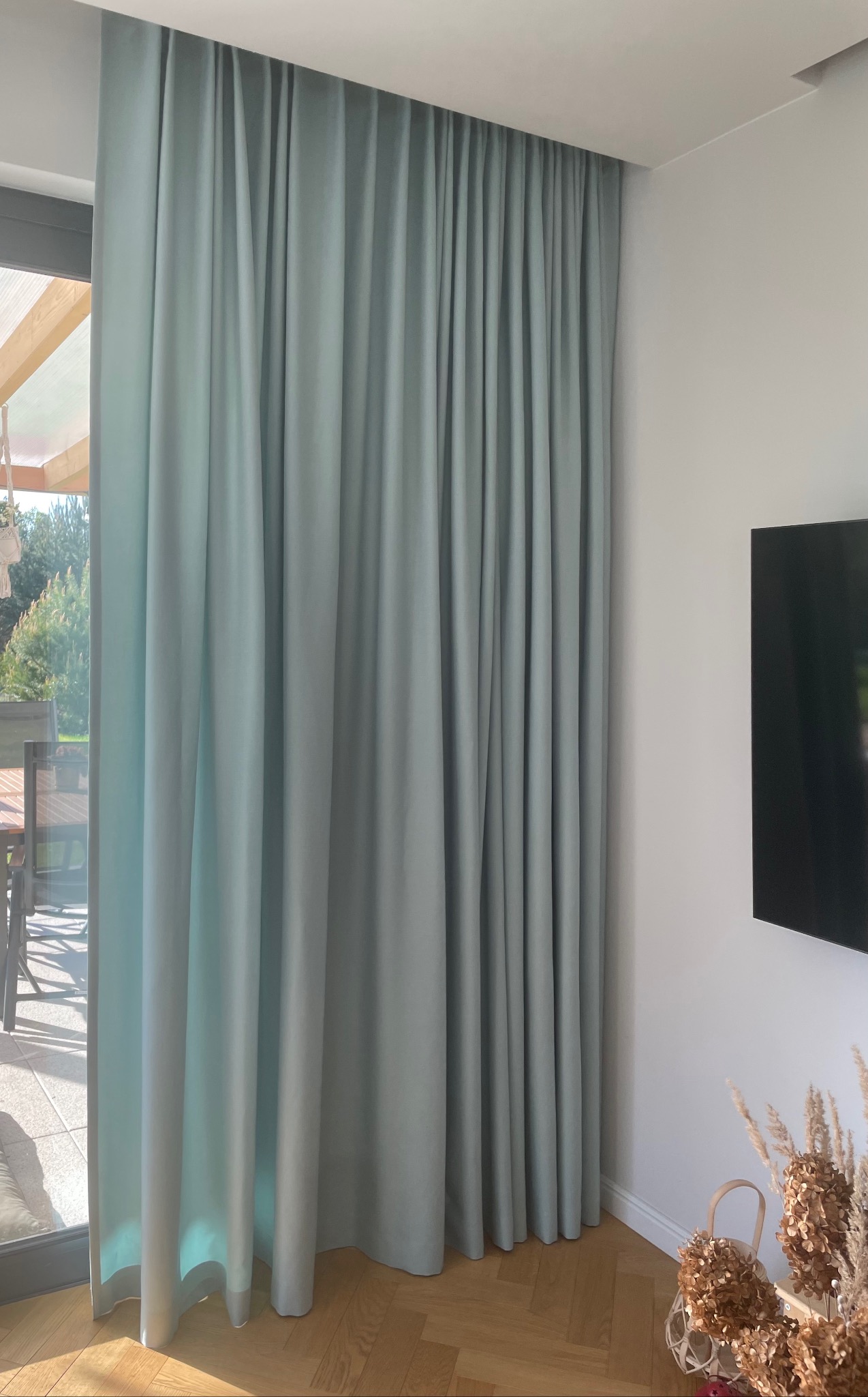 Długa, gładka zasłona w odcieniu pastelowej zieleni, zawieszona przy oknie balkonowym. Widoczny fragment podłogi z parkietu i telewizor na ścianie.