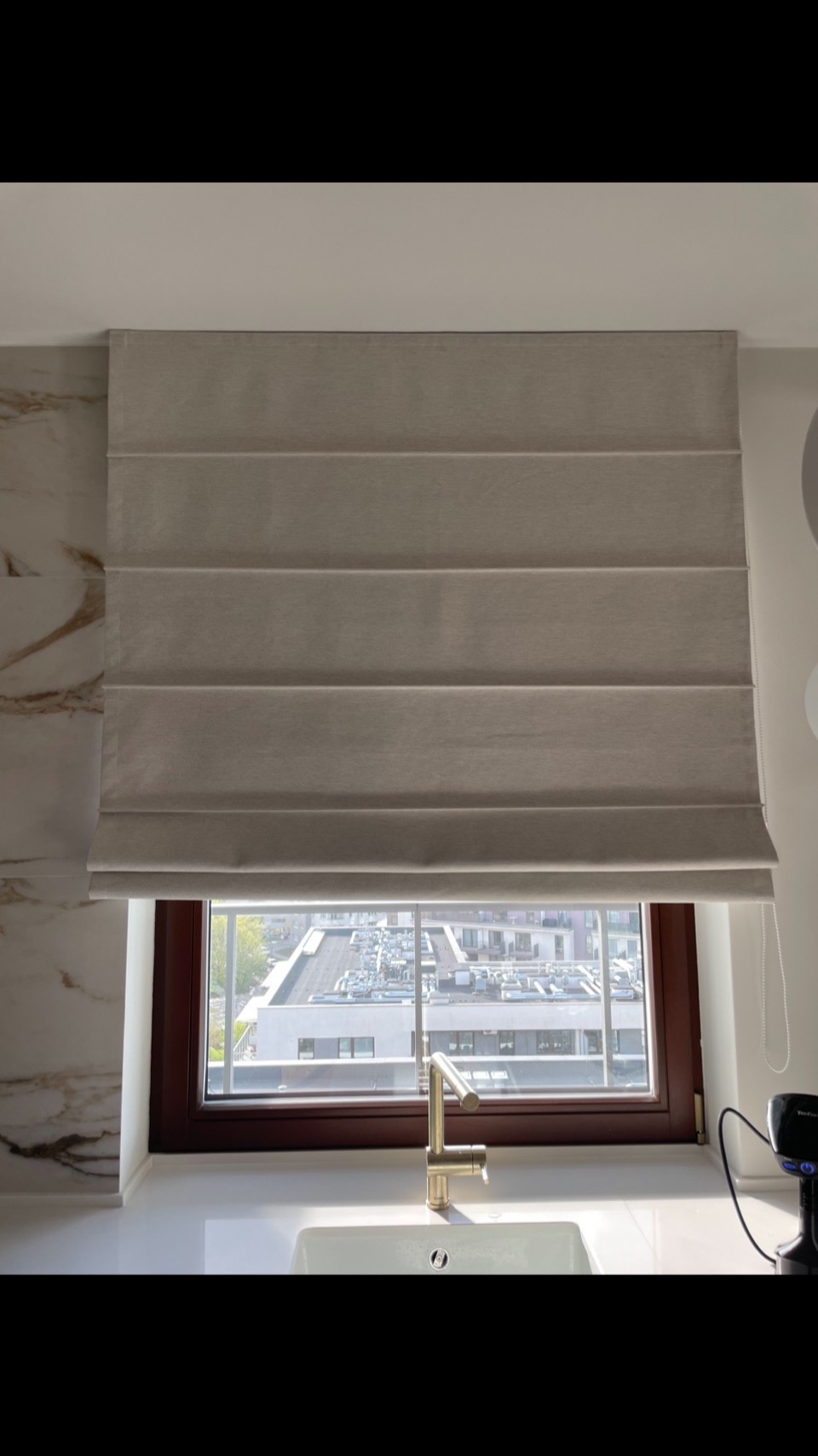 Beżowa roleta rzymska w oknie kuchni, z widokiem na budynki. Złota bateria przy zlewie, marmurowe wykończenie ściany po lewej stronie. Stonowana kolorystyka.