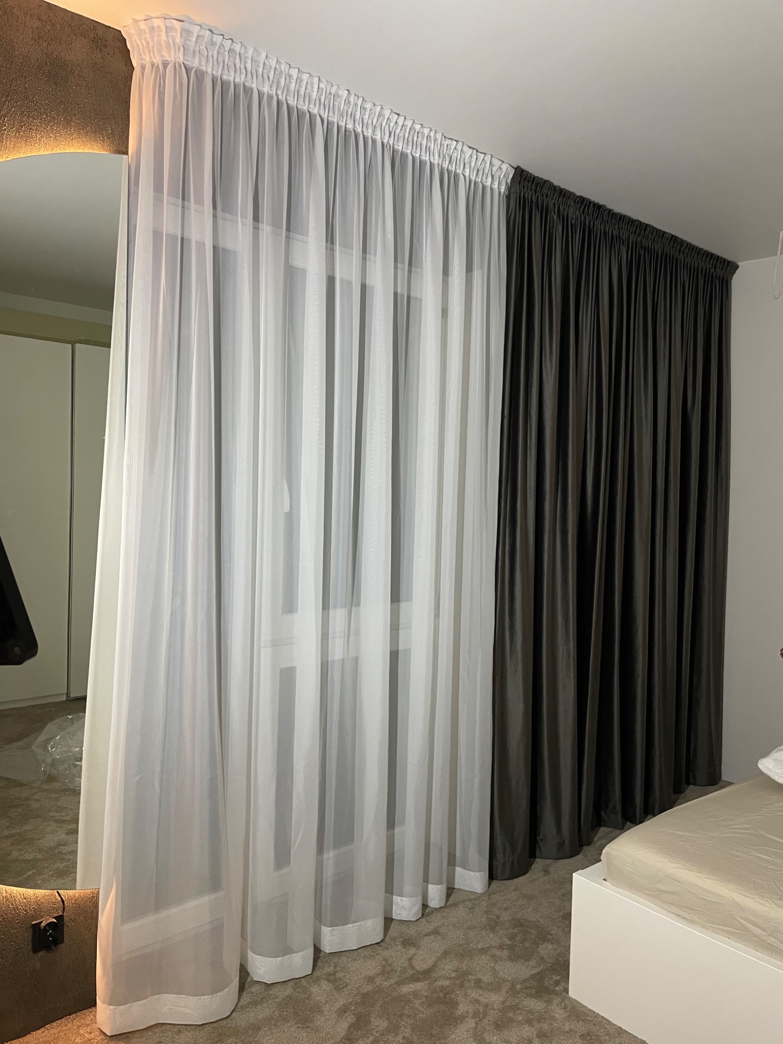 Okno z asymetrycznie dobranymi zasłonami: zwiewna biała firana i ciężka, grafitowa zasłona. Widoczny fragment sypialni z łóżkiem i szafą.