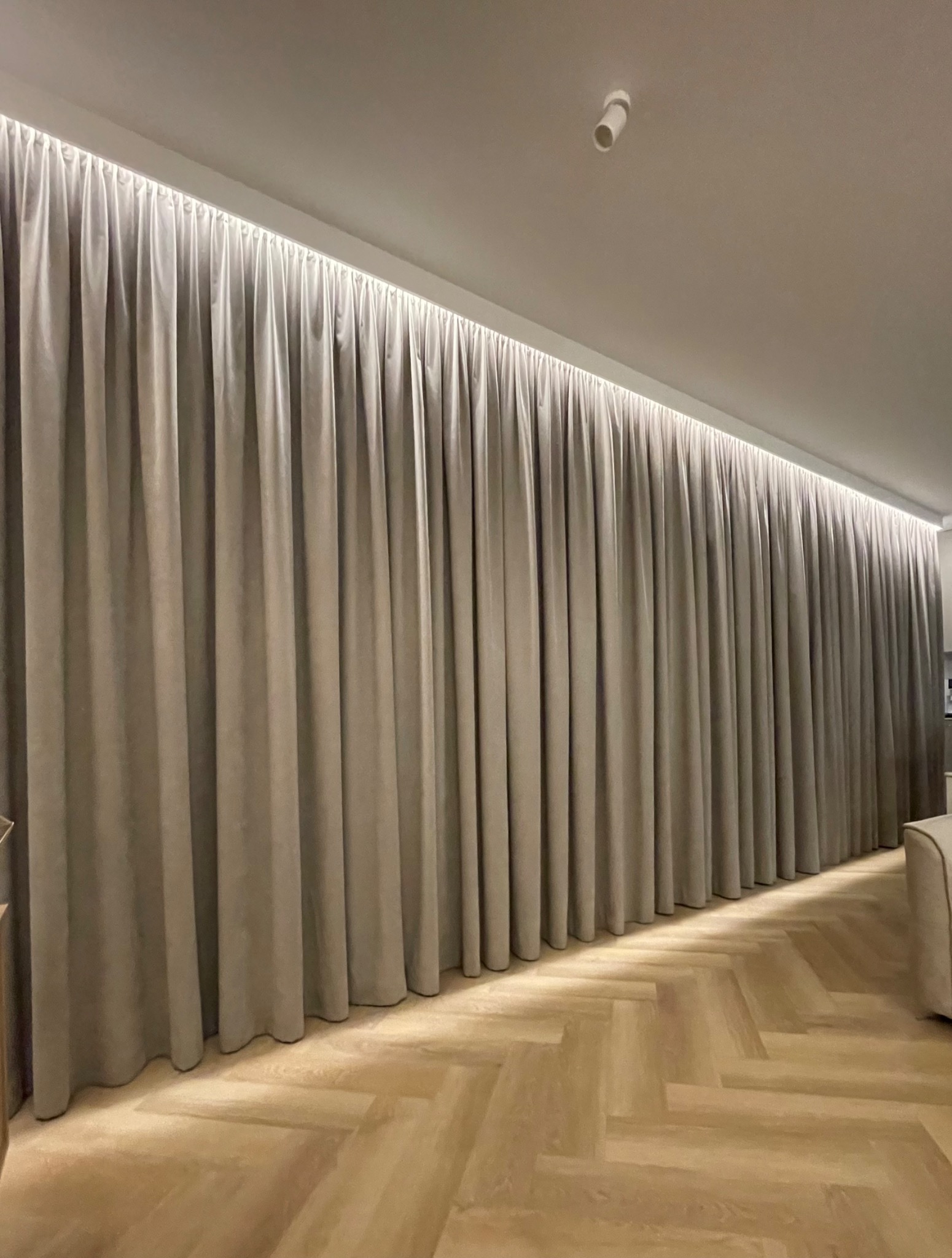 Beżowe, długie zasłony z delikatnego materiału, wiszące od sufitu do podłogi, oświetlone listwą LED, na tle parkietu ułożonego w jodełkę.