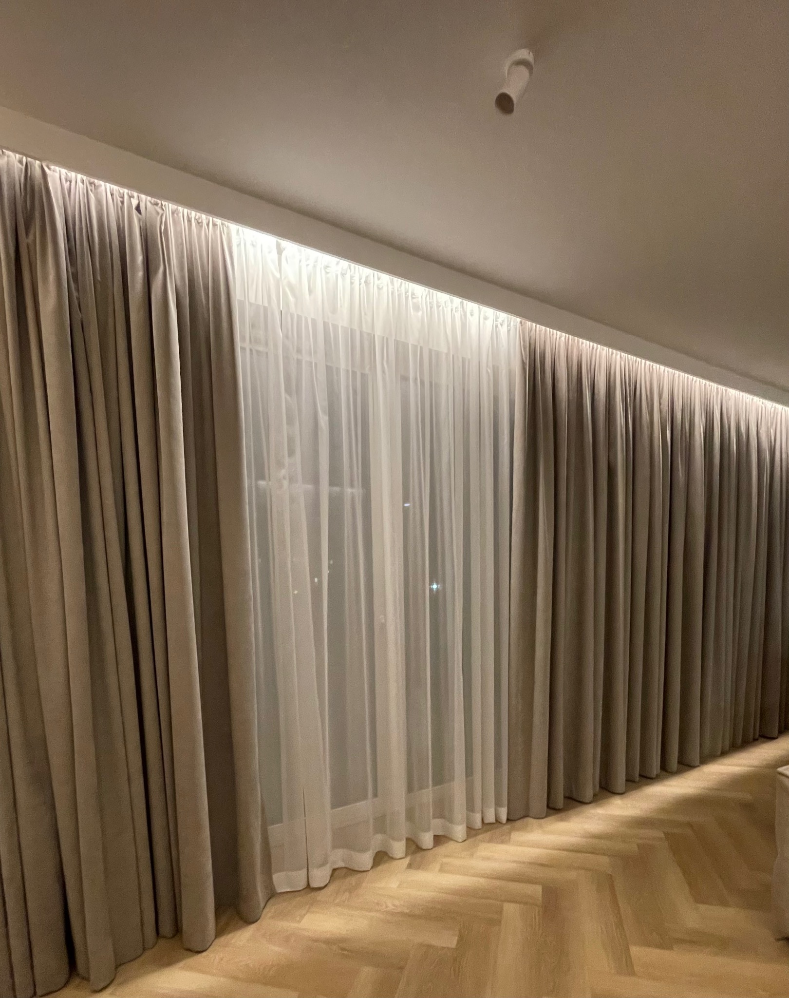 Beżowe, grube zasłony i białe, zwiewne firany, subtelnie oświetlone od góry, tworzą elegancką dekorację okienną w pomieszczeniu z parkietem w jodełkę.