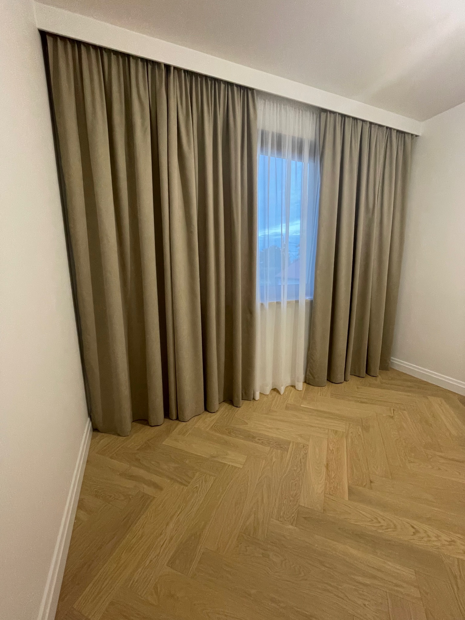 Beżowe zasłony zaciemniające z delikatną firanką w jasnym pokoju z parkietem ułożonym w jodełkę, widok na okno z fragmentem nieba.