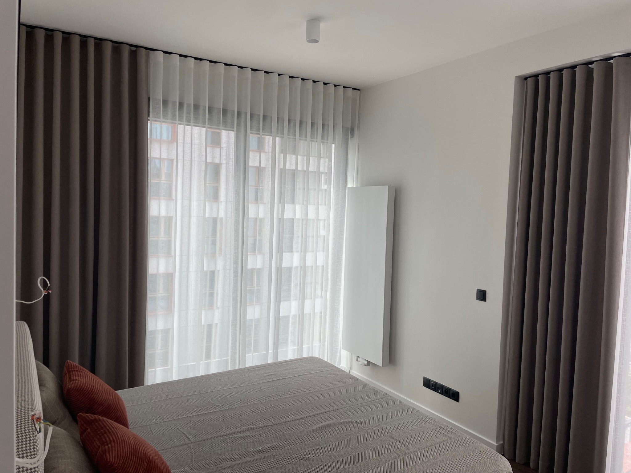 Nowoczesna sypialnia z zasłonami zaciemniającymi w odcieniach szarości i białymi, zwiewnymi firanami. Widok na budynek przez okno. Minimalistyczny wystrój.