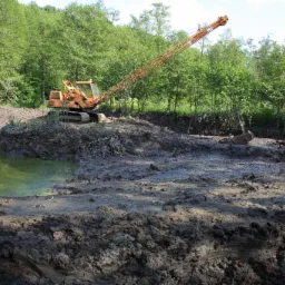Pomarańczowa koparka gąsienicowa z długim wysięgnikiem na brzegu wykopanego stawu, widoczne świeże ślady pracy w błotnistym terenie, w tle zielony las.