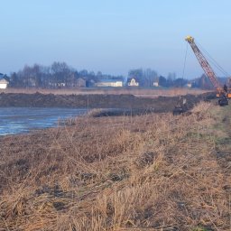STAW-BUD Sylwester Pawłoś - Żuraw gąsienicowy przy wykopanym stawie, w tle zabudowania wiejskie. Brzeg stawu z roślinnością, woda częściowo zamarznięta. Błękitne niebo.
