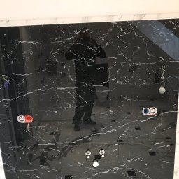 Dekor-Style - Ściana i podłoga wyłożone czarnym marmurem z białymi żyłami w trakcie prac wykończeniowych w łazience, widoczne puszki elektryczne, rury i odbicie osoby robiącej zdjęcie.