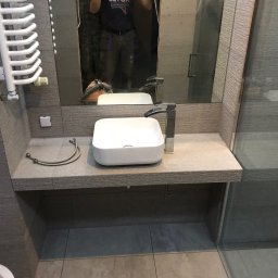 Dekor-Style - Nowoczesna łazienka w szarościach: biała, prostokątna umywalka nablatowa z chromowaną baterią, grzejnik drabinkowy i kabina prysznicowa ze szklanymi drzwiami. W lustrze odbicie osoby i odkurzacza.