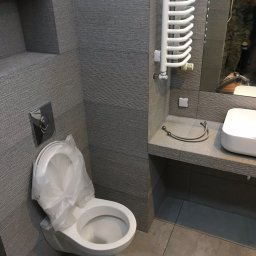 Dekor-Style - Szara łazienka w trakcie remontu: toaleta z założoną folią ochronną, umywalka na blacie, grzejnik drabinkowy i odbicie osoby w lustrze.