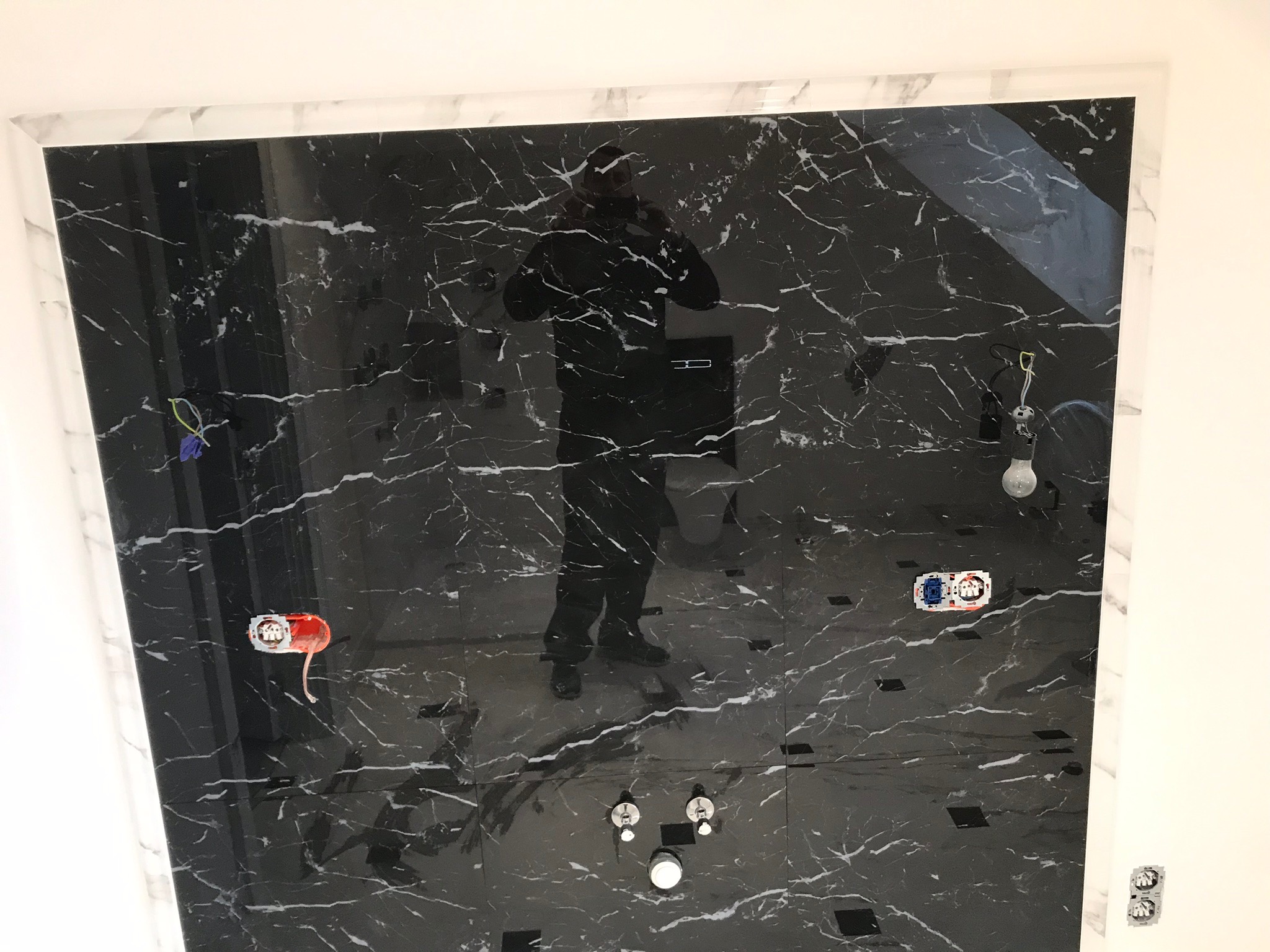 Ściana i podłoga wyłożone czarnym marmurem z białymi żyłami w trakcie prac wykończeniowych w łazience, widoczne puszki elektryczne, rury i odbicie osoby robiącej zdjęcie.