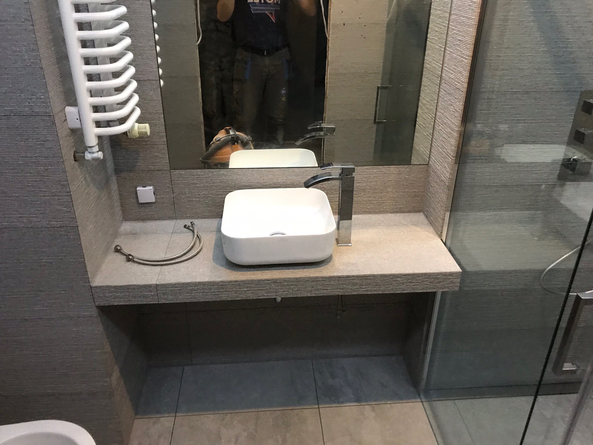 Nowoczesna łazienka w szarościach: biała, prostokątna umywalka nablatowa z chromowaną baterią, grzejnik drabinkowy i kabina prysznicowa ze szklanymi drzwiami. W lustrze odbicie osoby i odkurzacza.