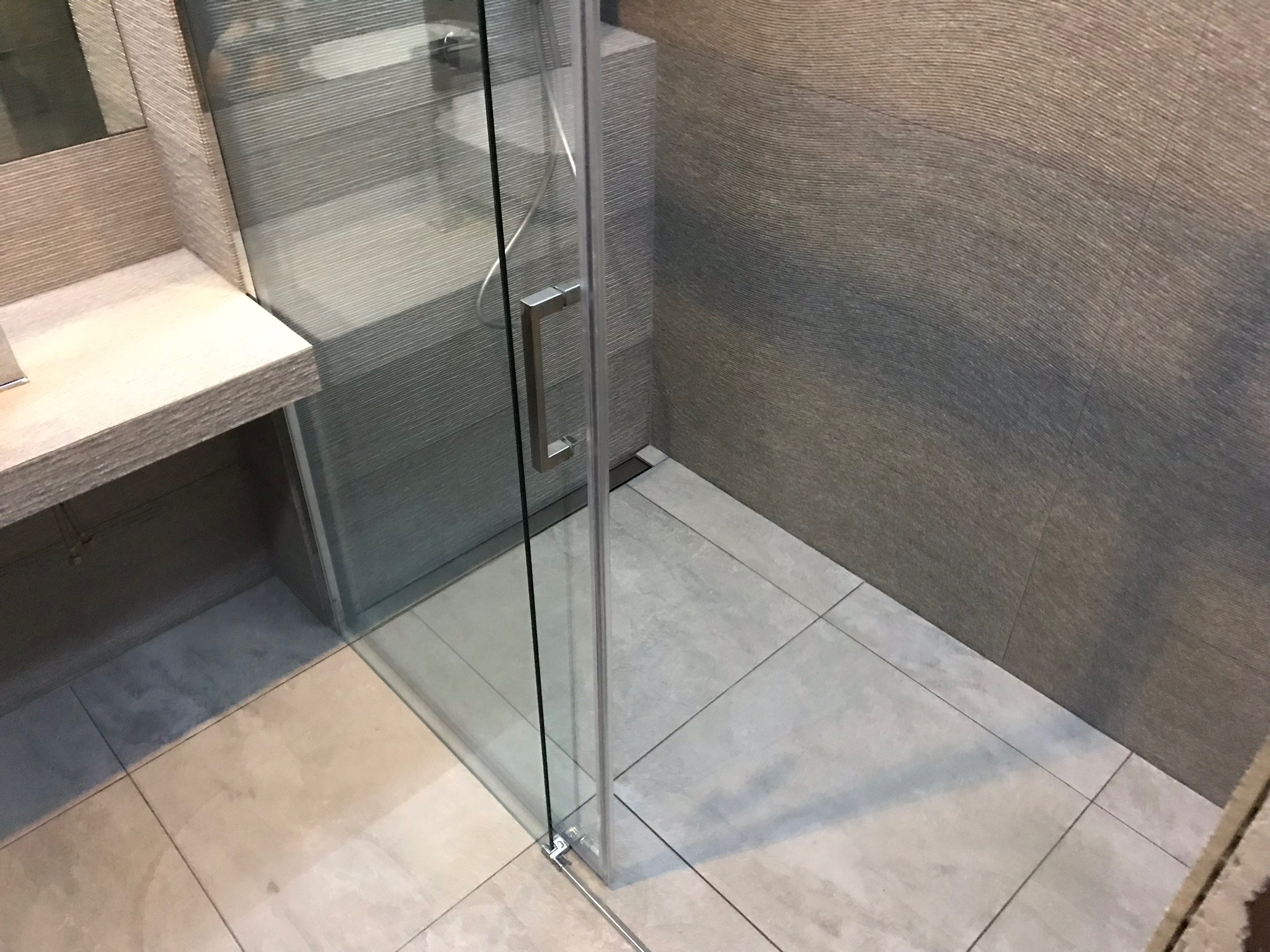 Szklana kabina prysznicowa z chromowanym uchwytem, jasne płytki podłogowe i ściany w odcieniach szarości z wyraźną fakturą, widok z góry.