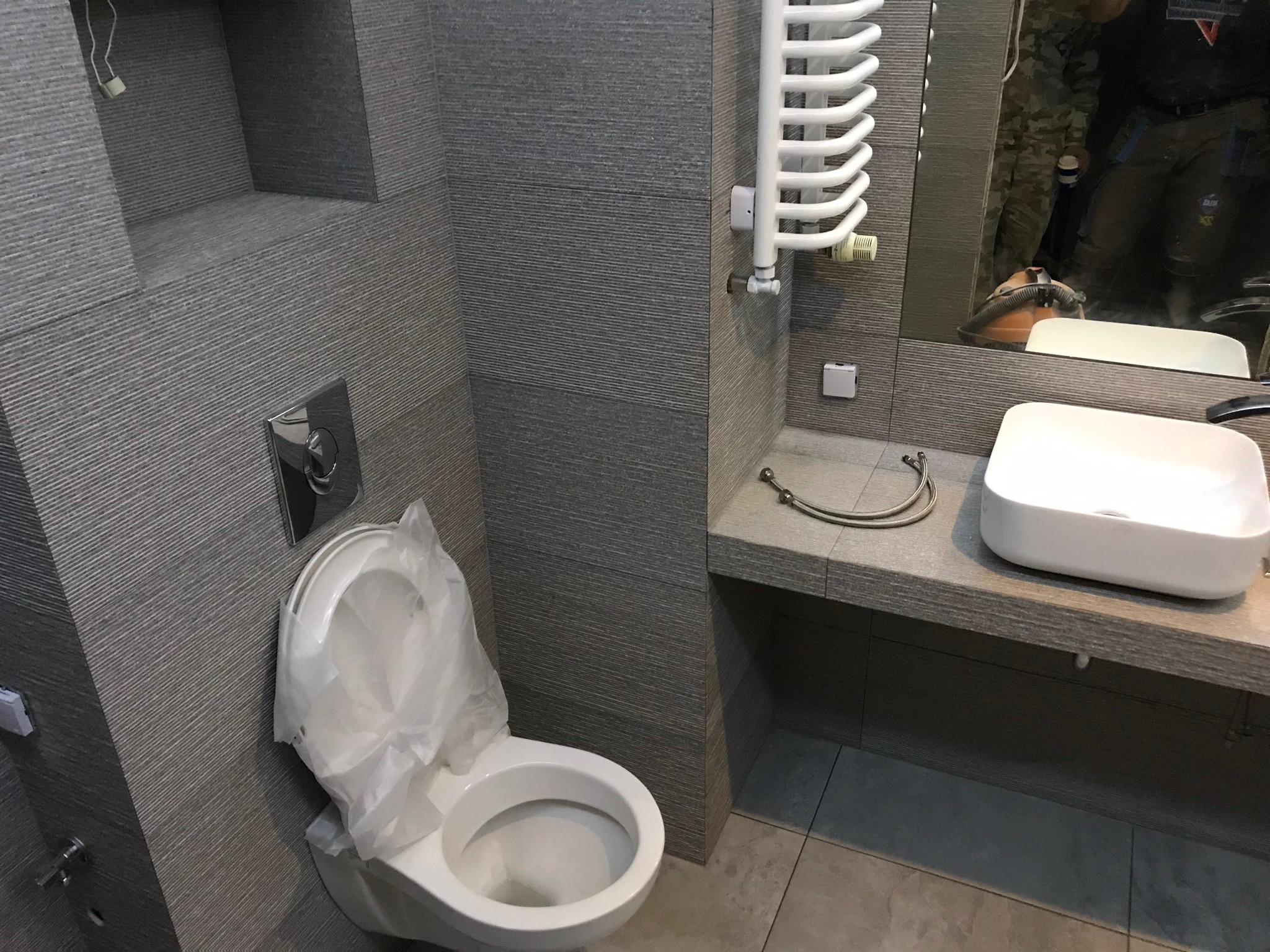 Szara łazienka w trakcie remontu: toaleta z założoną folią ochronną, umywalka na blacie, grzejnik drabinkowy i odbicie osoby w lustrze.