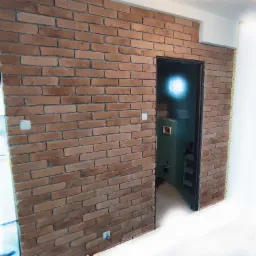 Ściana wykończona cegłą klinkierową w odcieniach brązu i beżu, widoczny fragment włącznika światła i przejście do innego pomieszczenia z zielonym regałem.