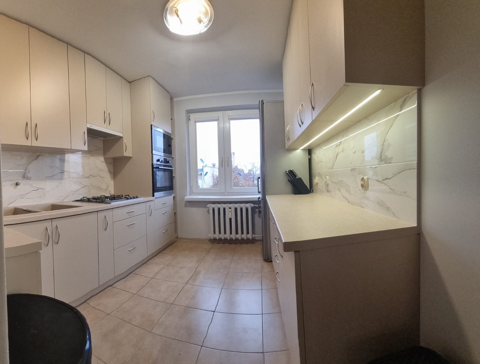 Jasna kuchnia z beżowymi szafkami, marmurowymi płytkami na ścianie i oświetleniem LED pod szafkami wiszącymi, widok na okno z grzejnikiem.