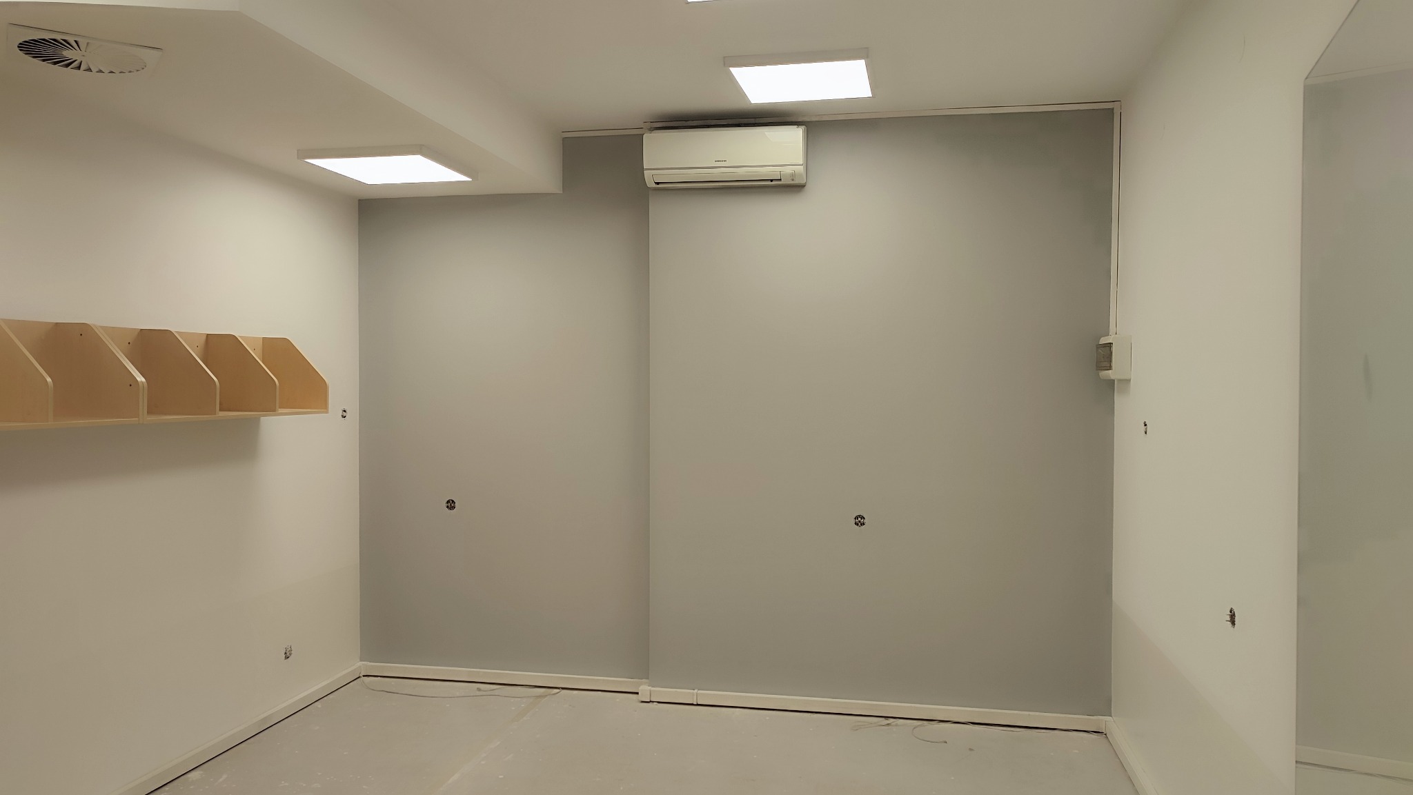 Wnętrze lokalu w trakcie remontu: pomalowane ściany w odcieniach szarości i bieli, zamontowany klimatyzator, widoczne puszki elektryczne i listwy przypodłogowe, surowa podłoga przygotowana...