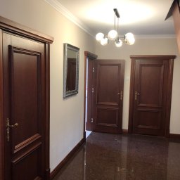 AdArt - Korytarz z trzema ciemnymi, drewnianymi drzwiami z ozdobnymi framugami, lustrem w srebrnej ramie i nowoczesną lampą sufitową z kulistymi kloszami.