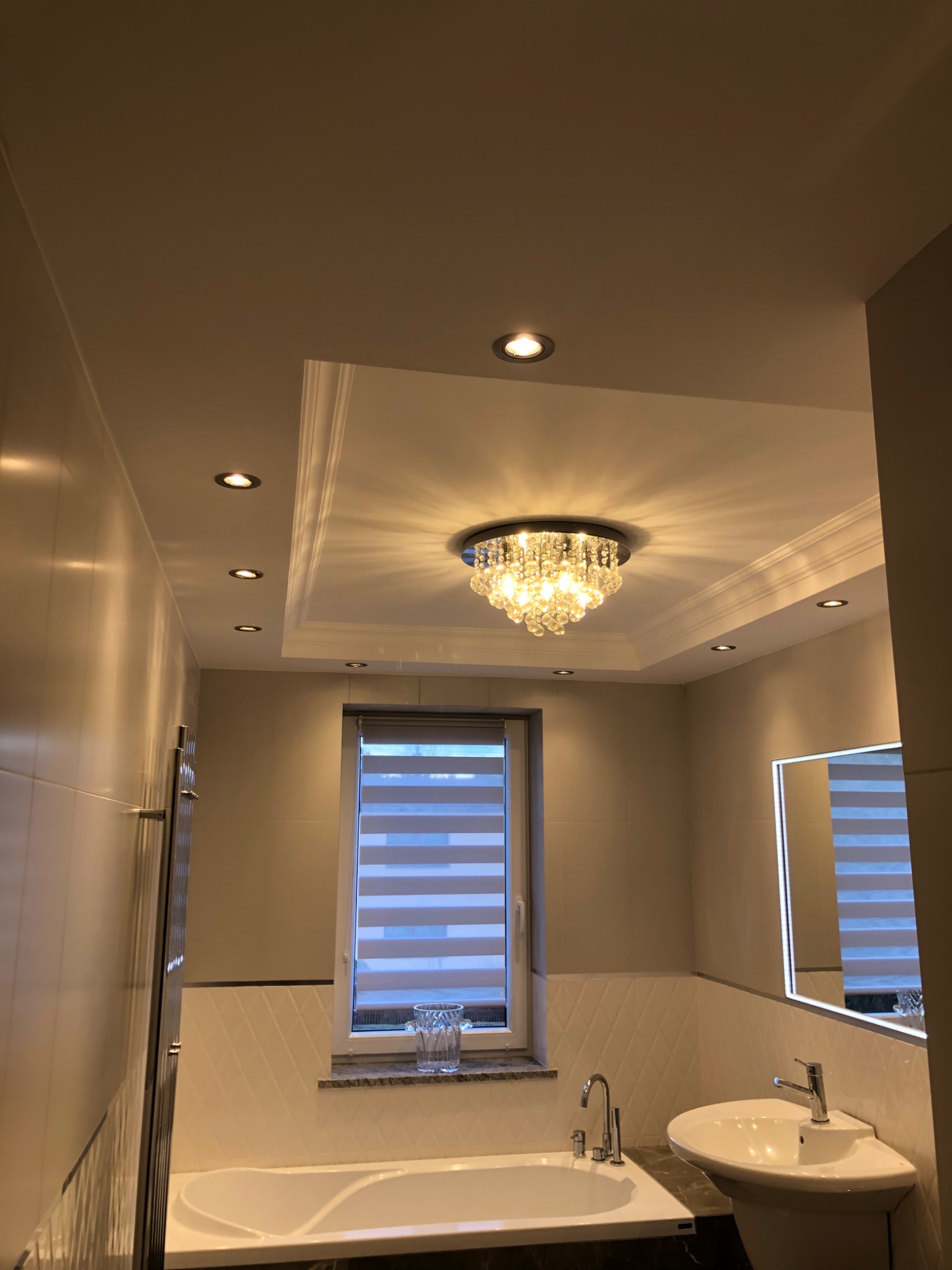 Elegancka łazienka z oświetleniem punktowym i kryształowym żyrandolem, białą armaturą oraz dekoracyjnymi płytkami ściennymi z motywem rombów.