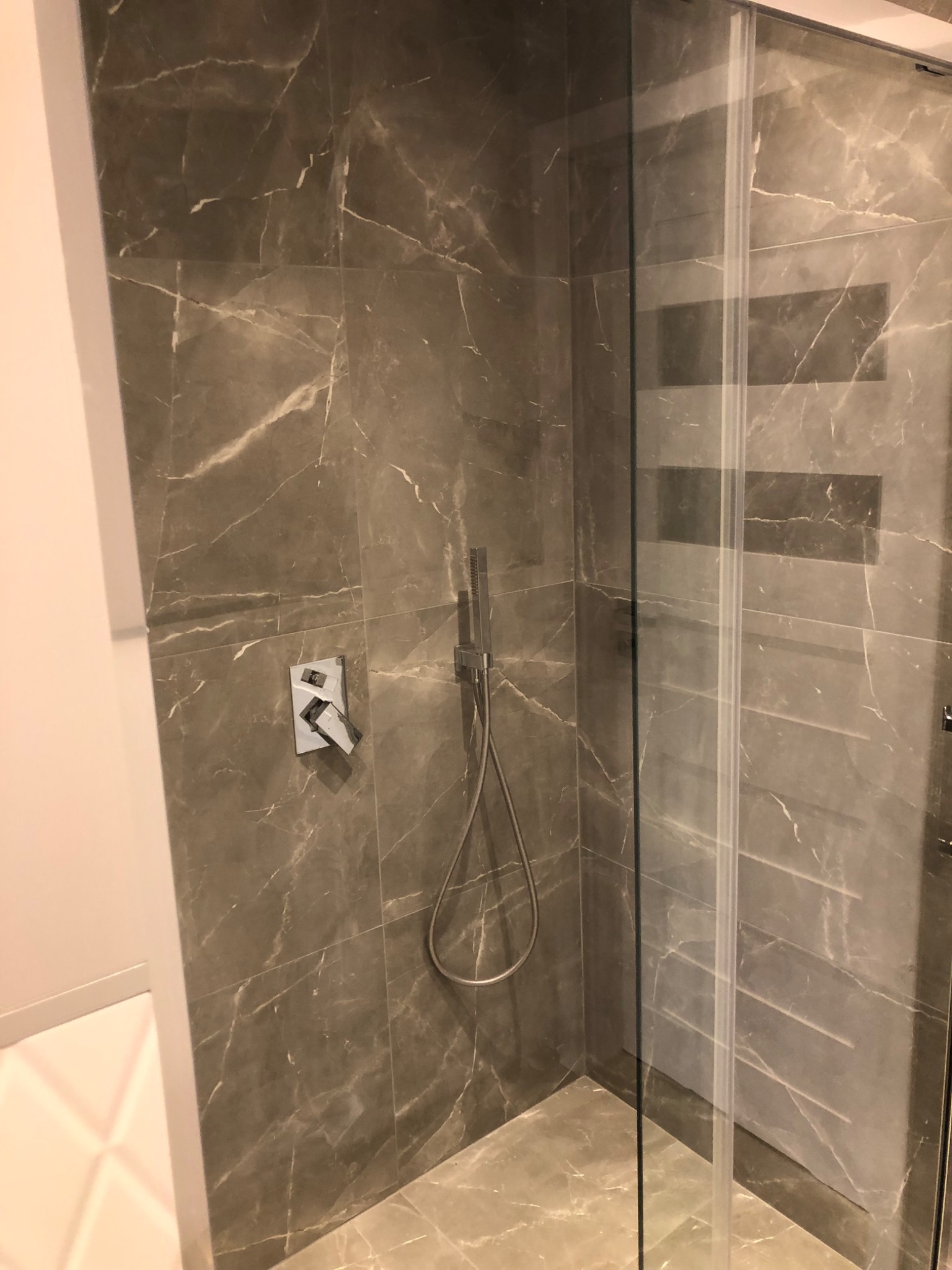 Szklana kabina prysznicowa z deszczownicą i baterią podtynkową, wykończona szarymi płytkami imitującymi marmur z wyraźnym wzorem żyłkowania na ścianach i podłodze.