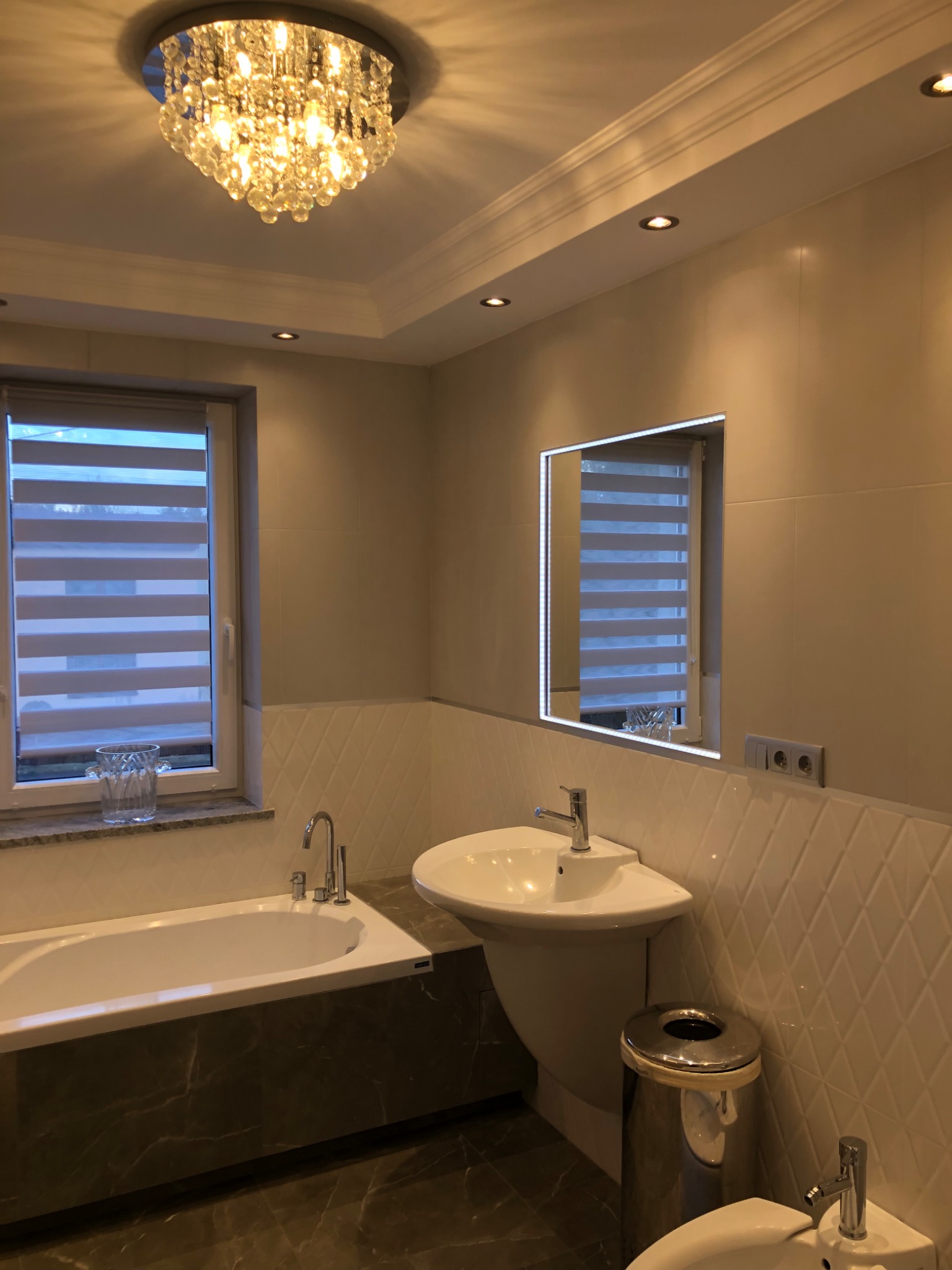 Elegancka łazienka z marmurową podłogą i obudową wanny, kryształowy żyrandol, podświetlane lustro, biała umywalka na postumencie i bidet.