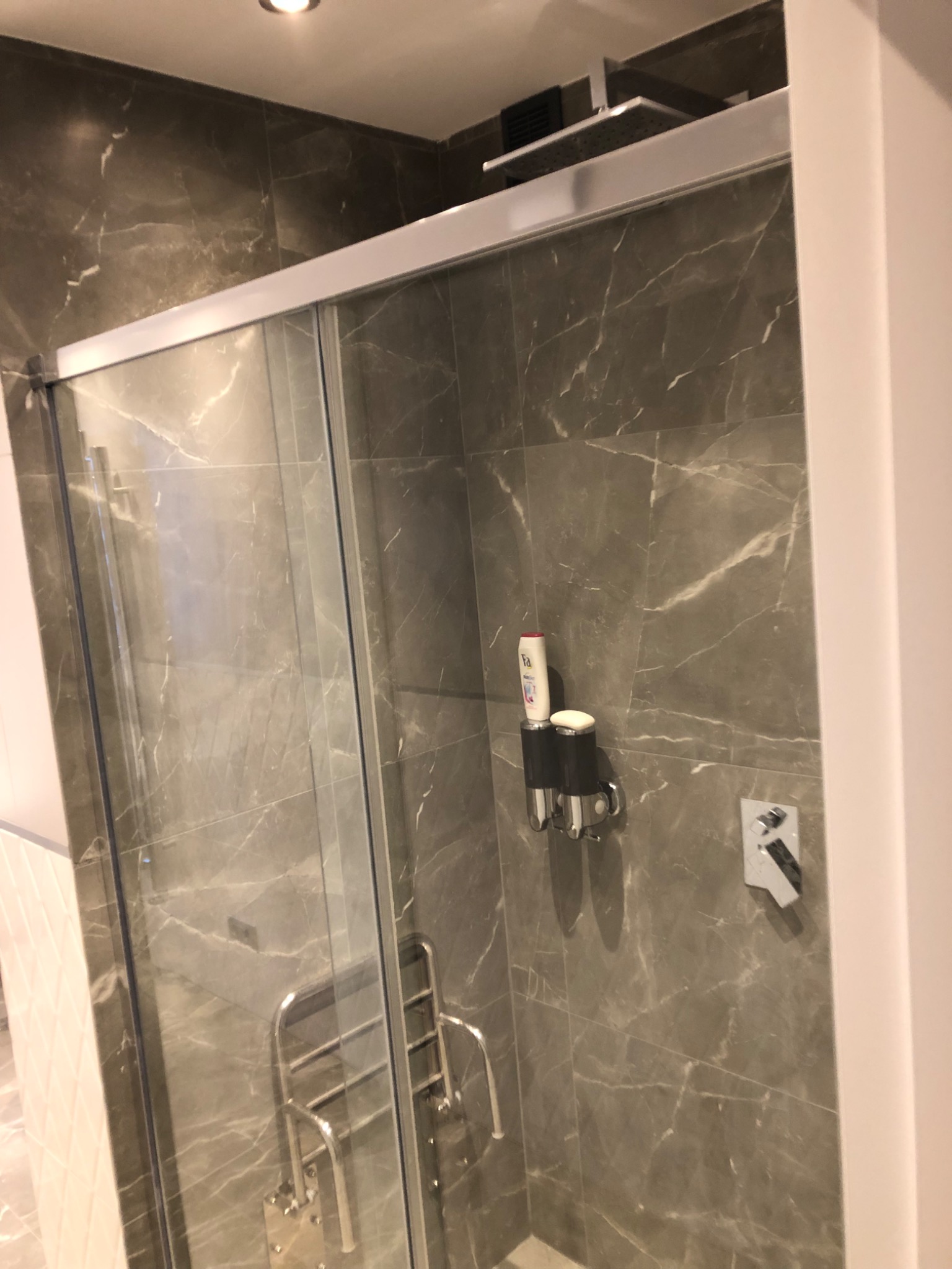 Kabina prysznicowa z przesuwnymi drzwiami, wykończona płytkami imitującymi marmur w odcieniach szarości, z zamontowanym dozownikiem na mydło i szampon oraz chromowanym grzejnikiem drabinkowym.