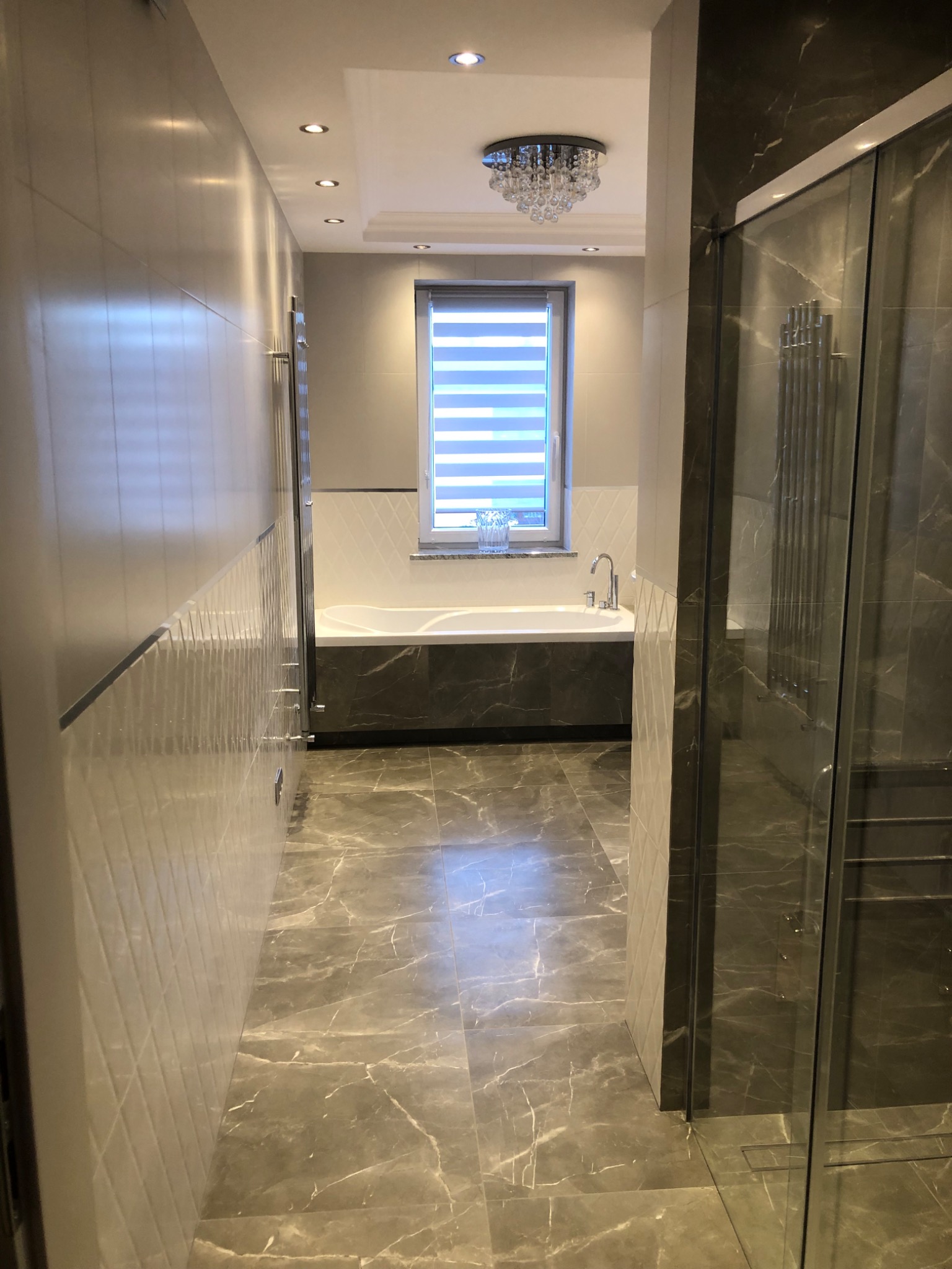 Elegancka łazienka w Krakowie z marmurową podłogą i ścianami, kabina prysznicowa ze szklanymi drzwiami, wanna pod oknem z żaluzjami poziomymi i ozdobny żyrandol kryształowy.