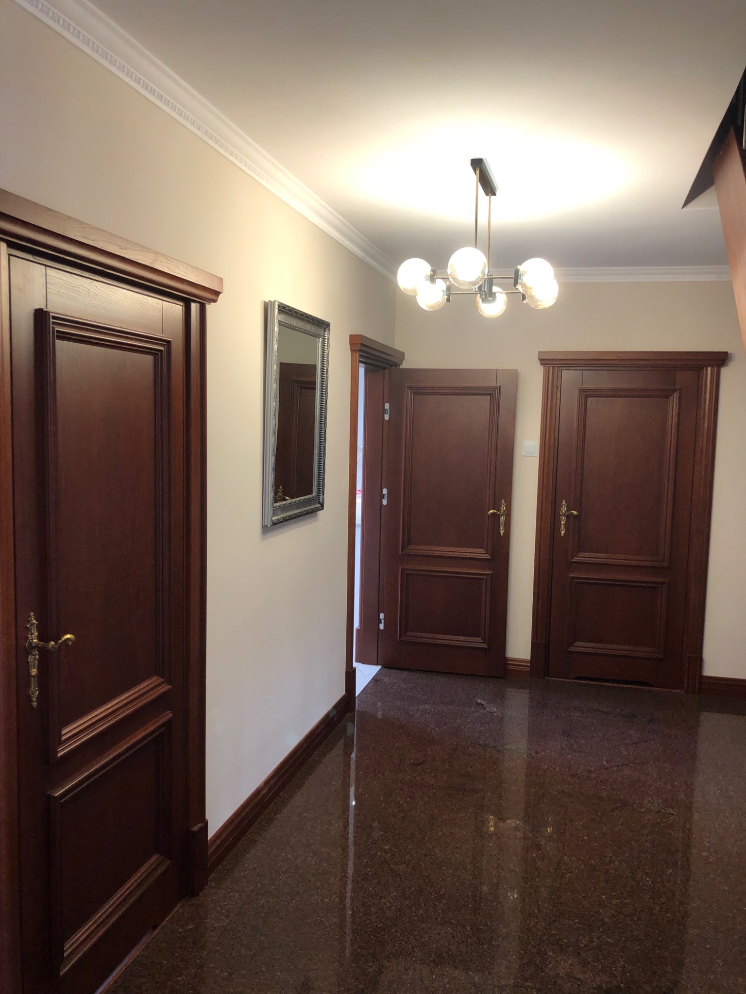 Korytarz z trzema ciemnymi, drewnianymi drzwiami z ozdobnymi framugami, lustrem w srebrnej ramie i nowoczesną lampą sufitową z kulistymi kloszami.