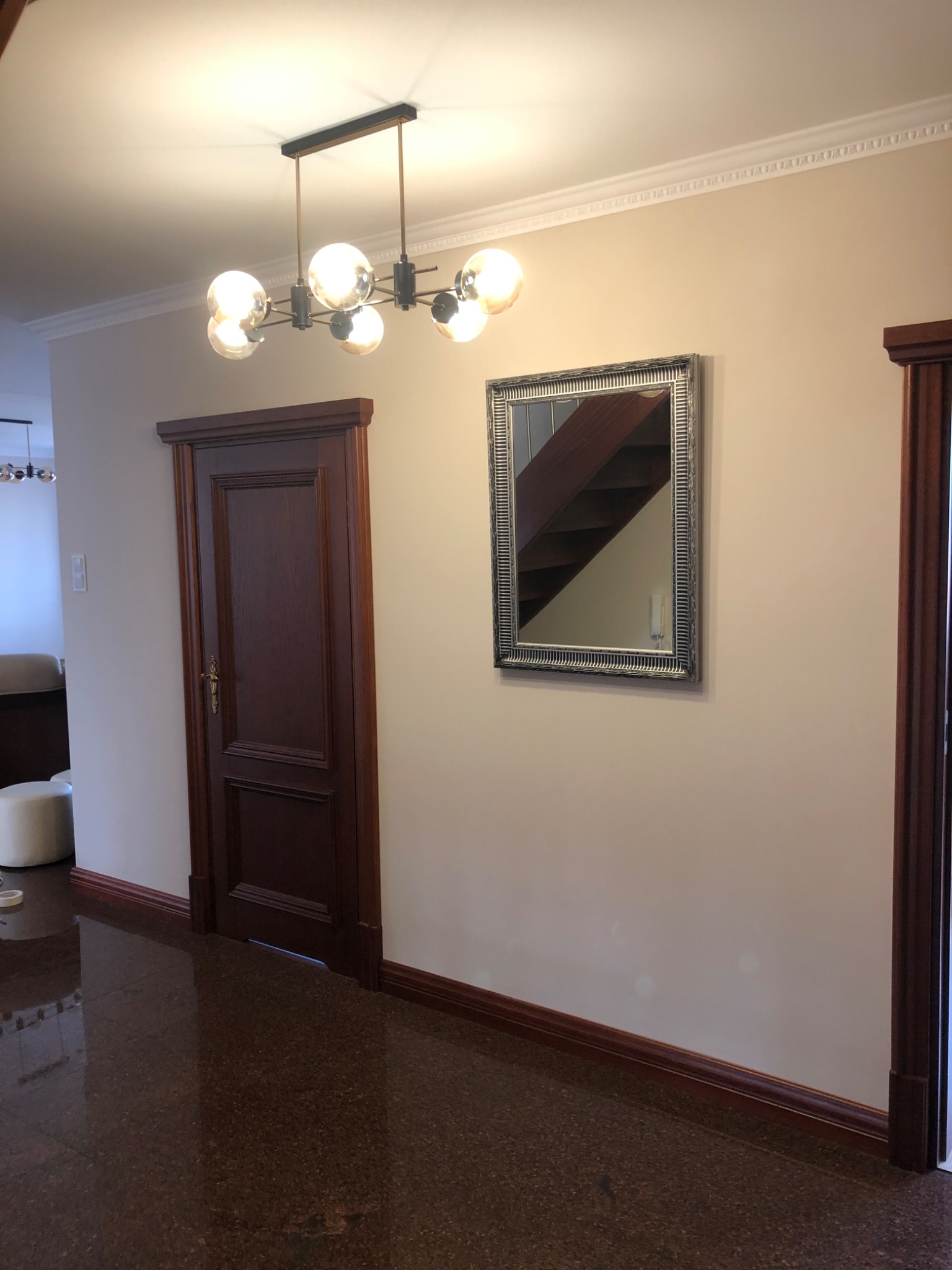 Wnętrze przedpokoju z ciemnobrązowymi drzwiami i listwami przypodłogowymi, lustro w srebrnej ramie odbija schody, nowoczesna lampa sufitowa z kulistymi kloszami, podłoga z terakoty o ciemnym odcieniu.