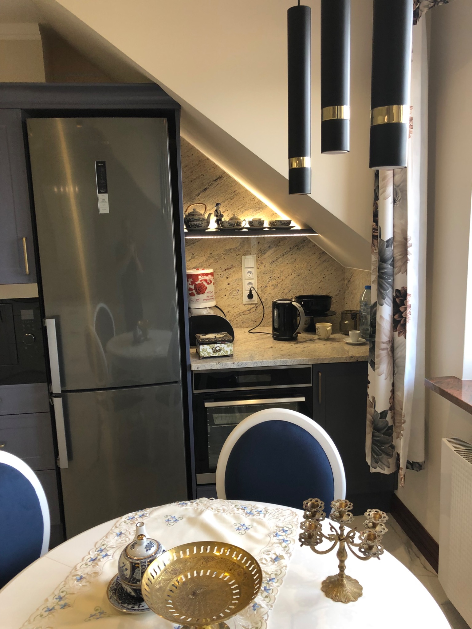 Nowoczesna kuchnia z szarymi szafkami, granitowym blatem i oświetleniem punktowym pod skośnym sufitem, uzupełniona o srebrną lodówkę i okrągły stół z dekoracyjnymi elementami.