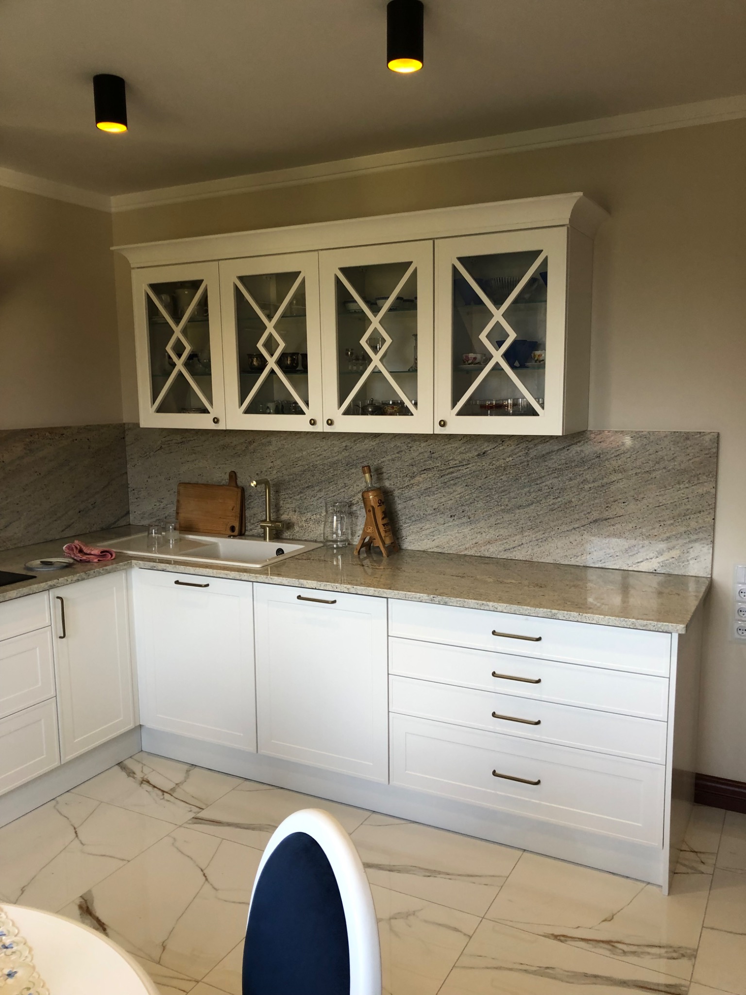 Jasna kuchnia z białymi szafkami, marmurowym blatem i backsplashiem, oraz ozdobnymi szklanymi frontami szafek wiszących, oświetlona czarnymi lampami sufitowymi.