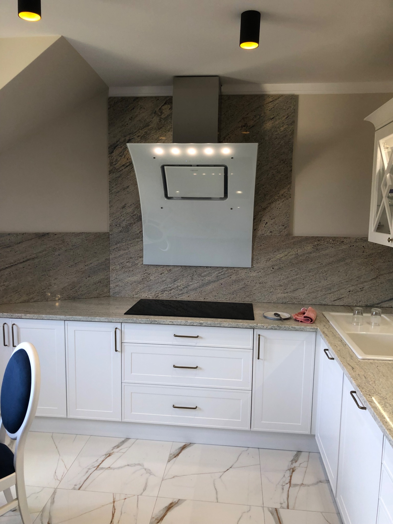 Nowoczesna kuchnia w odcieniach bieli i szarości, z marmurową podłogą i blatem, oraz białymi szafkami z ciemnymi uchwytami, oświetlona punktowymi lampami sufitowymi.