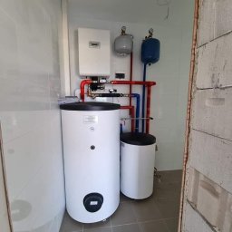 Sol Fin - Nowa instalacja grzewcza z dwoma białymi zbiornikami, kotłem i osprzętem w pomieszczeniu z szarymi płytkami i ścianami z białych płytek oraz surowego betonu.