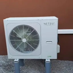 Zewnętrzna jednostka pompy ciepła marki Netsu zamontowana na szarych rurach PCV, na tle brązowej ściany i żwirowego podłoża.