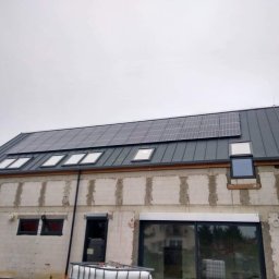 Sol Fin - Niedokończony budynek z szarego bloczka z zamontowanymi panelami słonecznymi na metalowym dachu i oknami dachowymi, widoczne zbiorniki na wodę i fragment samochodu dostawczego.