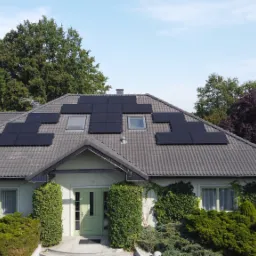 Dom jednorodzinny z szarym dachem, na którym zamontowano panele słoneczne w kilku sekcjach, z pracownikiem na dachu w tle.