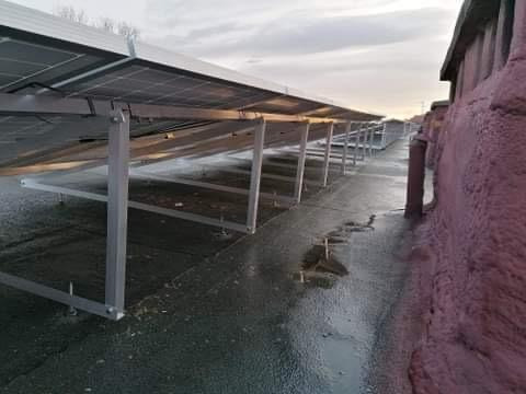 Rząd paneli słonecznych zamontowanych na płaskim dachu, widok z dołu, ukazujący system montażowy i ocieplenie pianką PUR na ścianie budynku w tle.