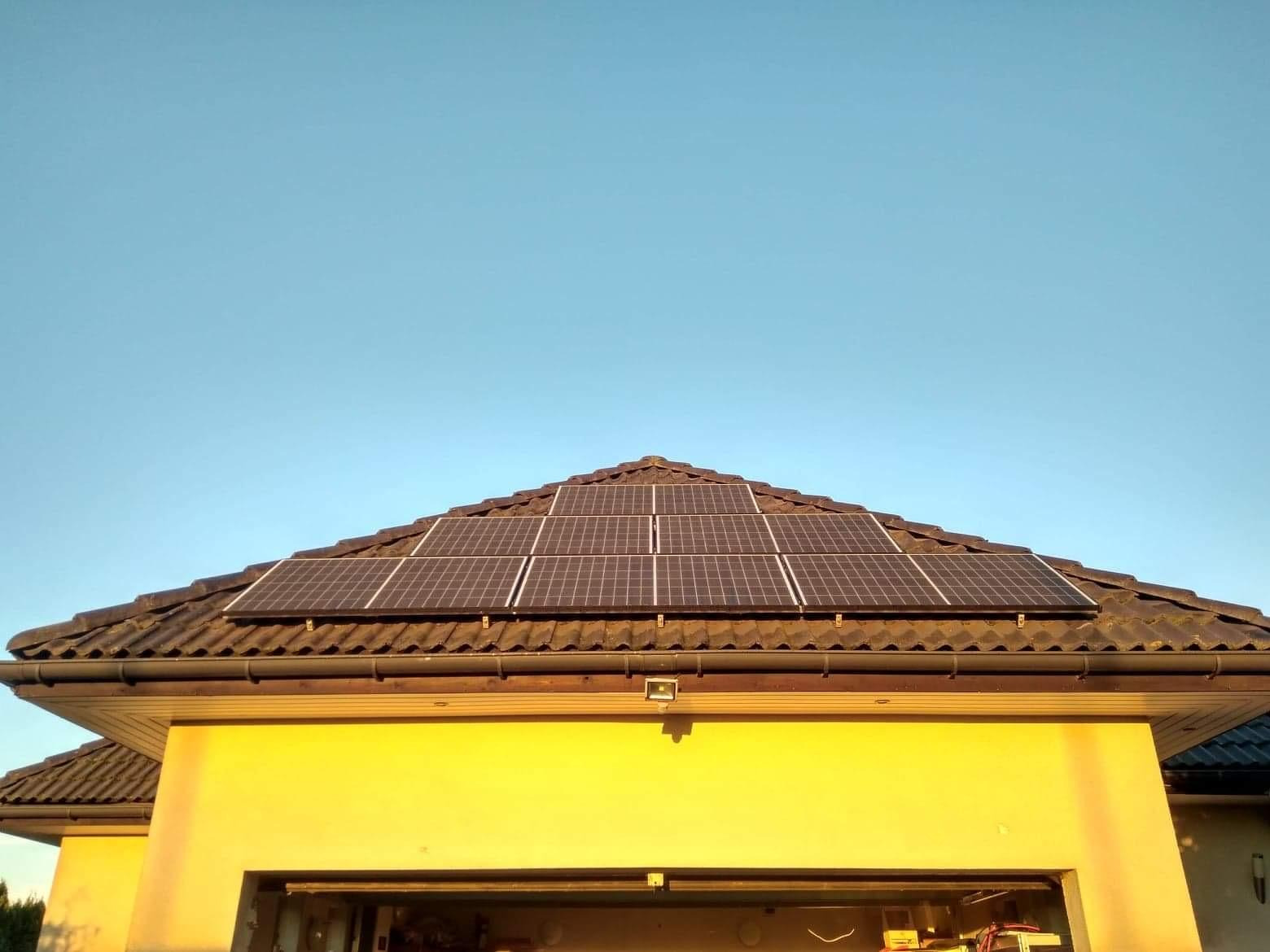 Panele słoneczne na brązowym dachu domu z żółtą elewacją, widok od dołu pod kątem, błękitne niebo w tle.