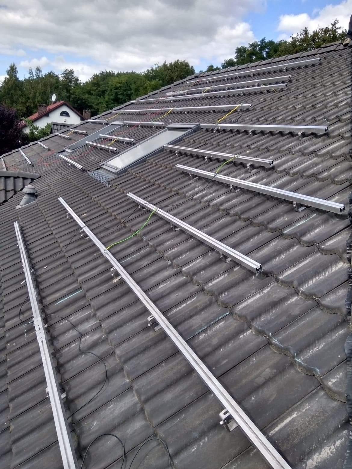 Montaż aluminiowych szyn pod panele słoneczne na dachu z dachówki ceramicznej, widoczne kable i elementy mocujące, w tle dom z oknem dachowym.