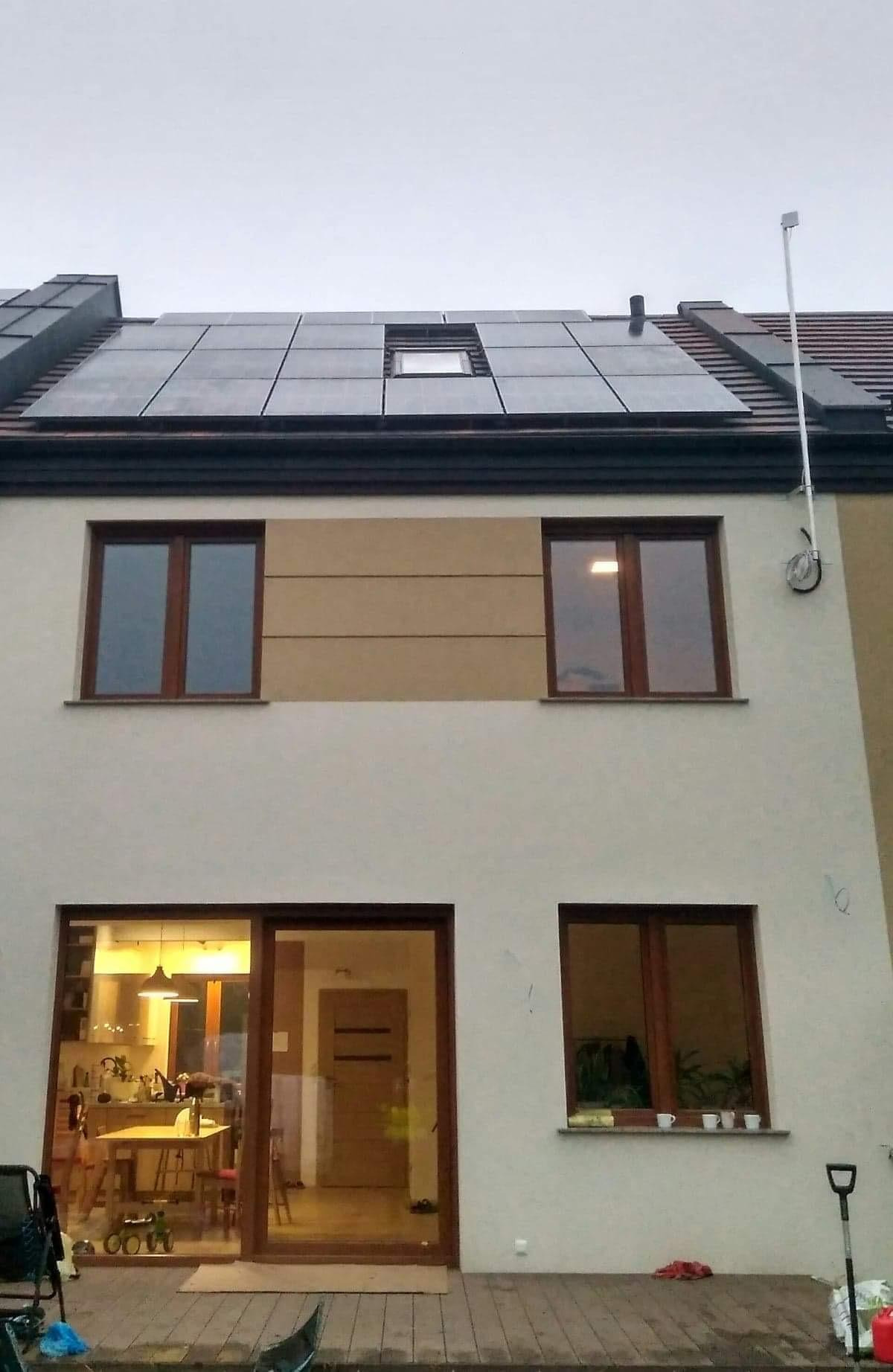 Dwupiętrowy dom z panelami słonecznymi na dachu, widoczne okno dachowe, elewacja w odcieniach bieli i beżu, z przodu taras z desek.