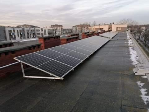 Instalacja fotowoltaiczna na płaskim dachu budynku z widocznymi panelami, aluminiową konstrukcją wsporczą i elementami dachu pokrytymi papą oraz fragmentami topniejącego śniegu.