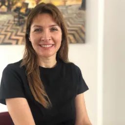 Portret uśmiechniętej kobiety z brązowymi włosami w czarnej koszulce na tle obrazu z geometrycznym wzorem.