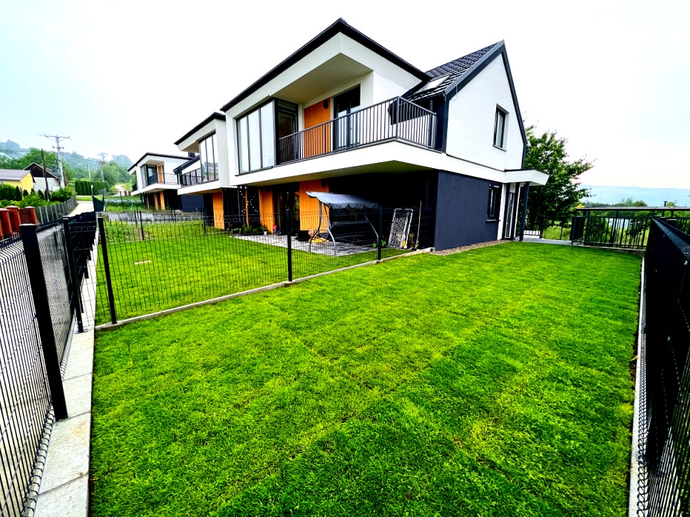 Nowoczesny dom szeregowy z czarną podmurówką, drewnianymi elementami elewacji i zadbanym trawnikiem za czarnym ogrodzeniem panelowym.