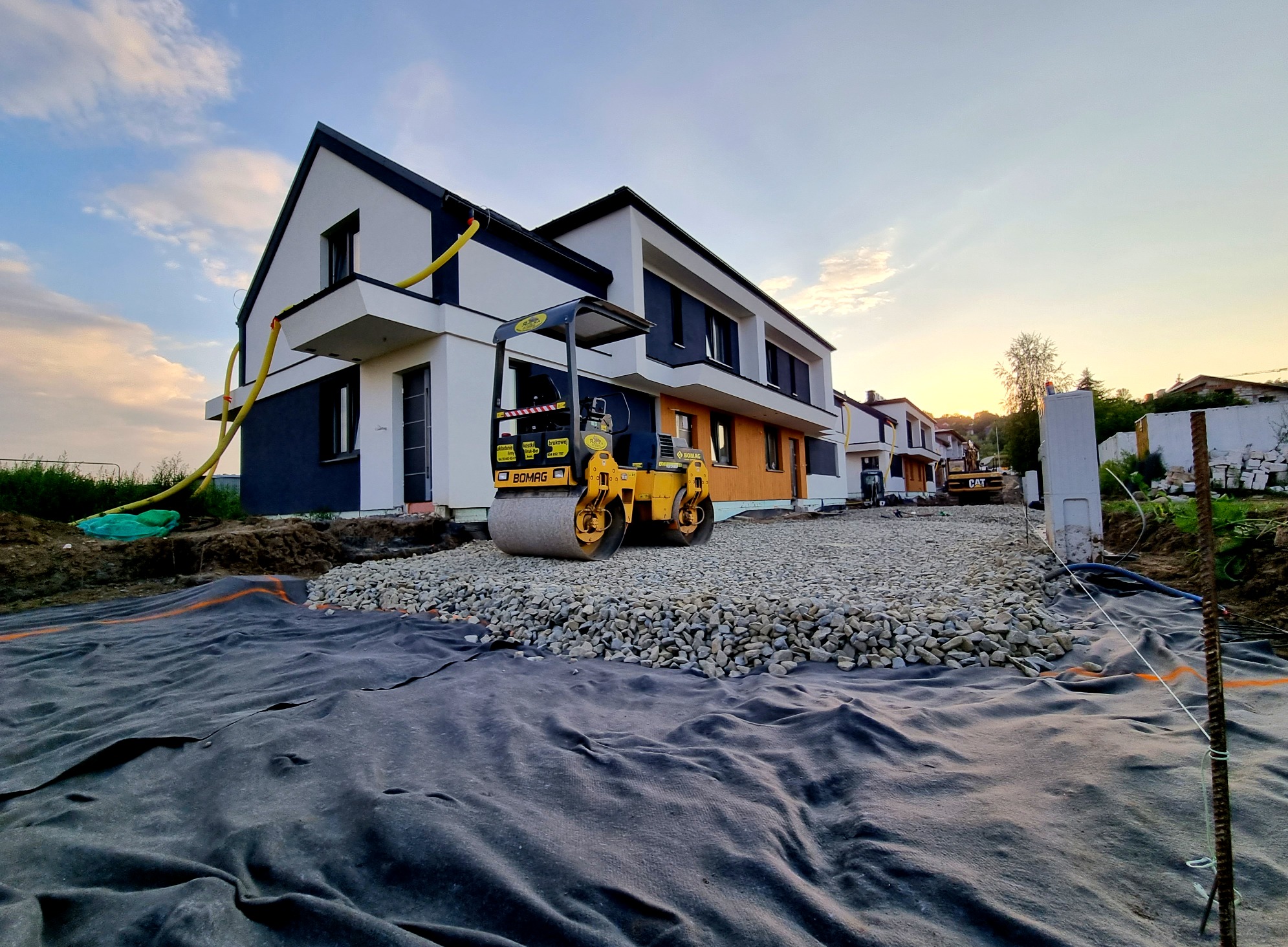 Przygotowanie terenu pod budowę domu w Nowym Sączu: żółty walec drogowy Bomag ubija kamienny podkład na czarnej geowłókninie, w tle nowoczesne domy w zabudowie szeregowej i koparka.