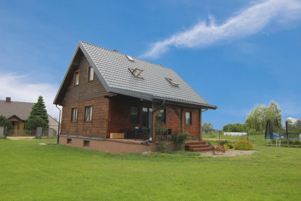 Drewniany dom z szarą dachówką i oknami dachowymi, otoczony zielonym trawnikiem, z widocznym trampoliną w tle i częścią sąsiedniego domu po lewej stronie.