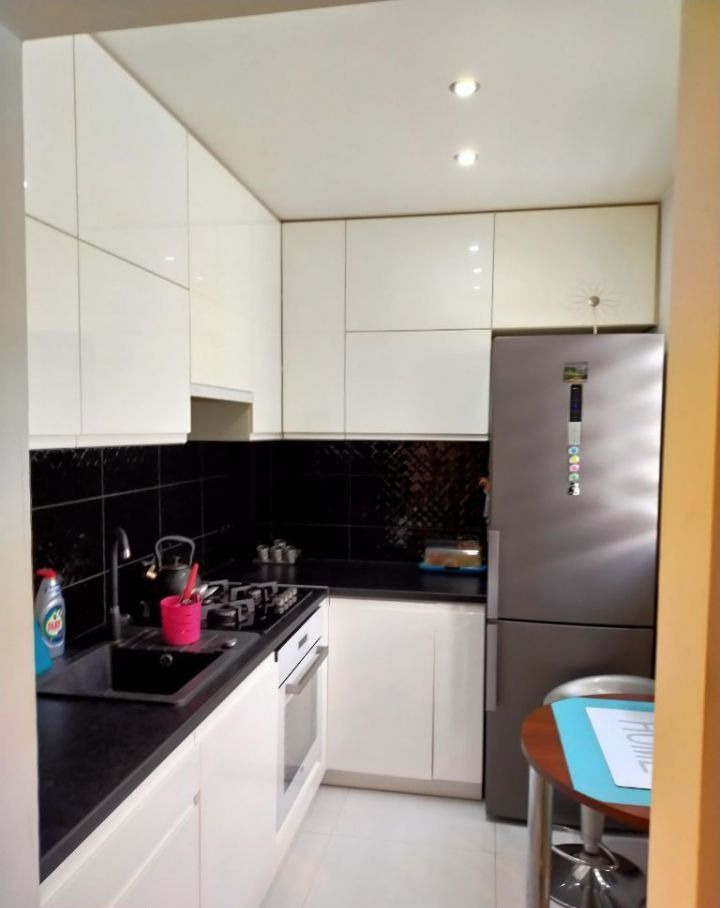 Wnętrze kawalerki: kompaktowa kuchnia z białymi, błyszczącymi szafkami, czarnym blatem i płytkami, srebrną lodówką oraz okrągłym stolikiem barowym z niebieską podkładką, widok z perspektywy wejścia.