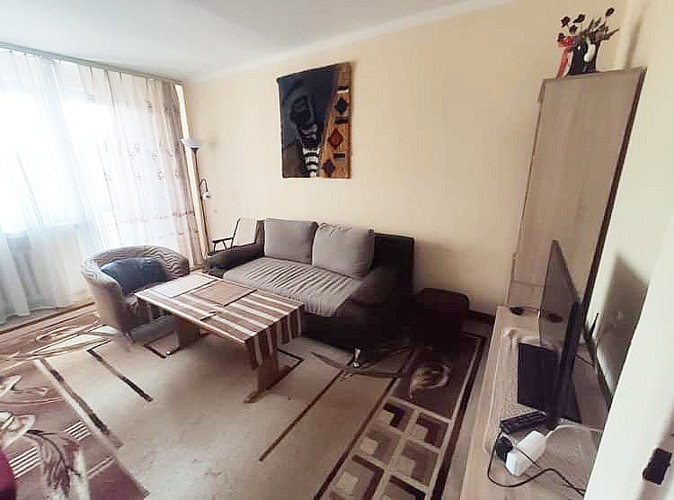 Jasny pokój dzienny z beżowymi ścianami, ciemnobrązową sofą z szarymi poduszkami, stolikiem kawowym w paski, wzorzystym dywanem i afrykańską maską na ścianie.