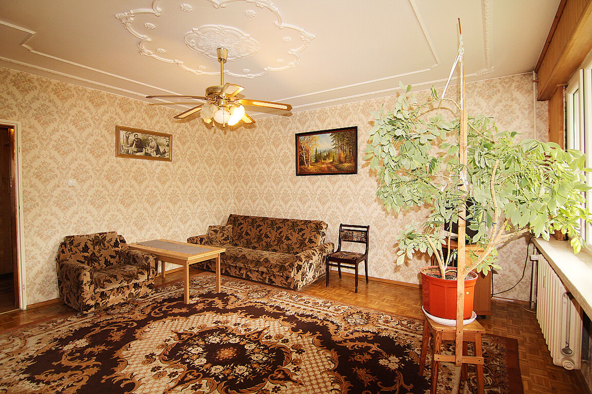 Umeblowane trzypokojowe mieszkanie z brązowym dywanem w kwiaty, wzorzystą tapetą, sofą i fotelem w podobnym stylu, oraz dużą rośliną doniczkową podpartą drewnianą konstrukcją.