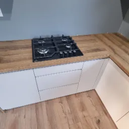 Kuchenna zabudowa z białymi frontami i drewnianym blatem, na którym umieszczono czarną płytę gazową Electrolux. Widok z góry, parkiet na podłodze.