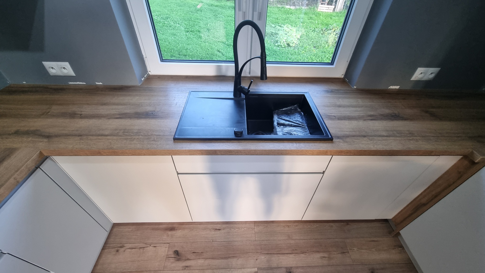 Czarny, minimalistyczny zlew z baterią na drewnianym blacie w kuchni pod oknem. Widoczne białe, gładkie fronty szafek i panele podłogowe. Nowoczesny design.