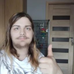 Osoba z uniesionym kciukiem na tle klatki z papugą, drzwi i szafy.