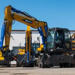 Żółte koparki SANY SY155W z niebieskimi osłonami na siłownikach, ustawione w rzędzie na placu, z żółtym samochodem serwisowym w tle.